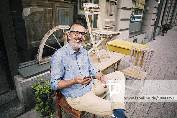 Portrait of owner holding mobile phone while sitting outside antique shop