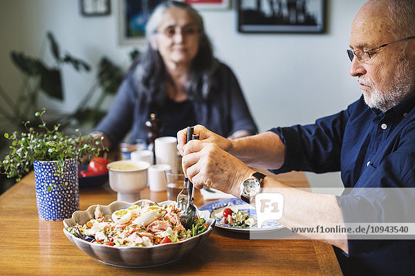Senior Mann serviert Salat für sich selbst  während er mit einer Frau am Tisch sitzt.
