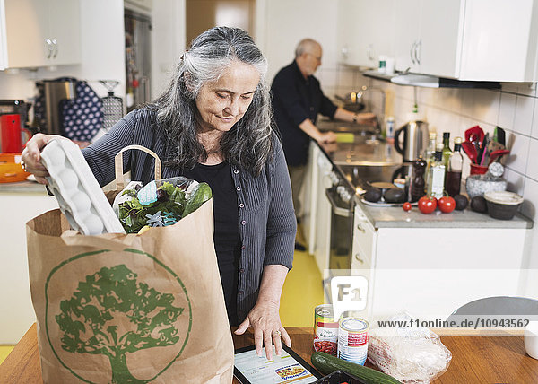 Senior Frau,  die Lebensmittel aus der Einkaufstasche nimmt,  während sie in der Küche ein digitales Tablett benutzt.