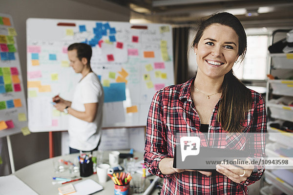 Porträt einer lächelnden Geschäftsfrau mit digitalem Tablett und Kollegin im Hintergrund
