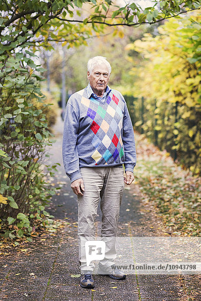 Ganzflächiges Porträt eines älteren Mannes  der auf einem Fußweg im Park steht.
