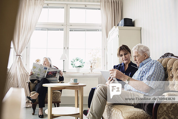 Senior man and caretaker using digital tablet while woman reading newspaper in nursing home