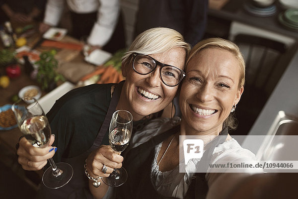 Cheerful mature women holding champagne flutes while taking selfie in kitchen