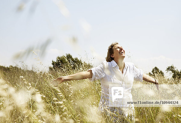 Fröhliche reife Frau lächelt beim Gehen mit erhobenen Armen im Grasfeld