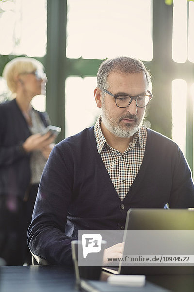 Mature businessman working on laptop while female coworker standing in office
