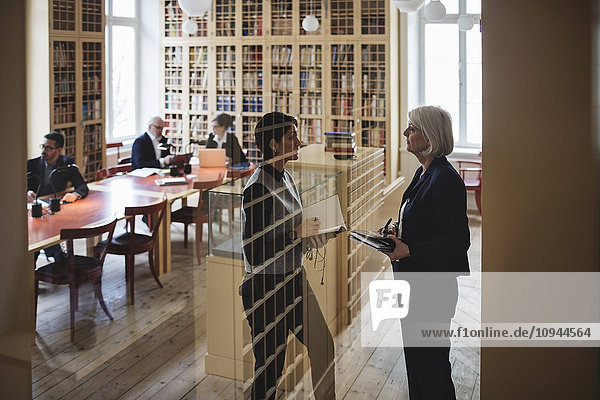 Female lawyers discussing while standing seen through glass in library