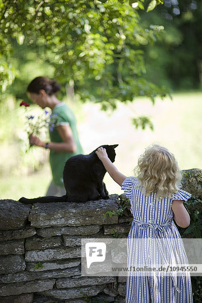 Mädchen streichelt Katze an der Wand  mit Mutter im Hintergrund