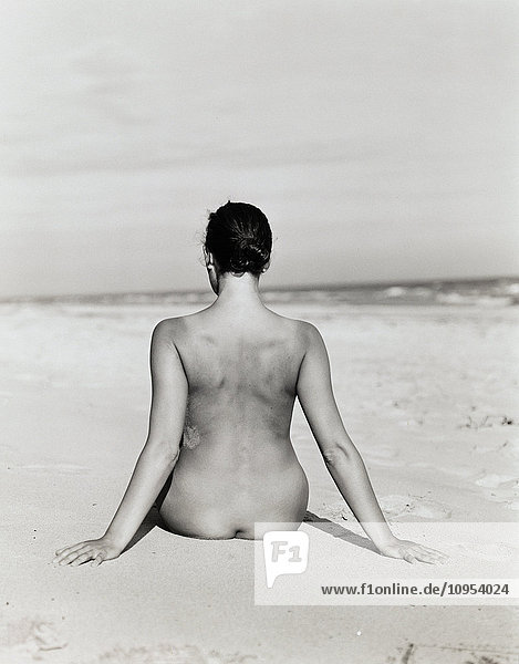 Nackte Frau sitzt am Strand mit dem Rücken zu.