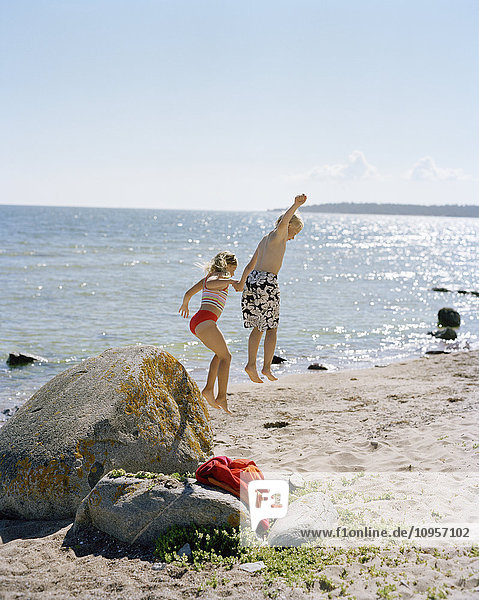 Junge und Mädchen springen von einem Stein am Strand  Schweden.
