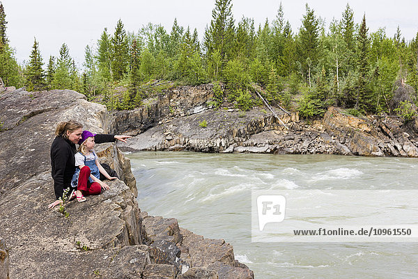 Mutter und Tochter sitzen auf einem Felsen am Flussufer und betrachten den Liard River  Whirlpool Canyon  British Columbia  Kanada  Sommer