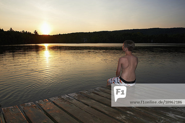 'Boy sitting on the dock with feet in water at sunset; Lac des Neiges  Quebec  Canada'