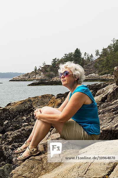 Frau sitzt auf einem Felsen am Ufer und blickt auf die Küste; Bowen Island  British Columbia  Kanada'.