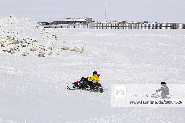 Einheimischer Alaska-Jugendlicher auf einer Schneemaschine  der einen anderen auf einem Schlitten zieht  die Stadt Barrow im Hintergrund  North Slope  Arktisches Alaska  USA  Winter'