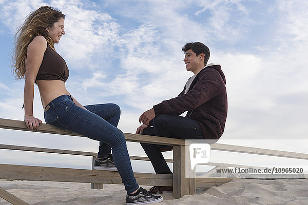 'A young man and woman sit talking on a wooden fence at the beach; Tarifa  Cadiz  Andalusia  Spain'