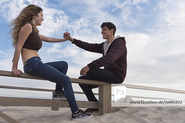 'A young man and woman sit talking and holding hands on a wooden fence at the beach; Tarifa  Cadiz  Andalusia  Spain'
