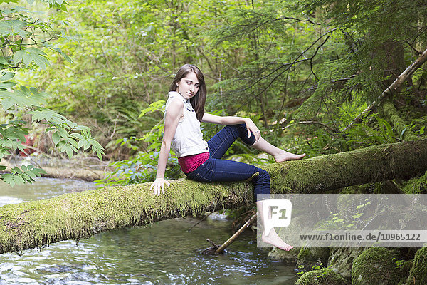 Porträt eines Mädchens im Teenageralter  das auf einem moosbewachsenen Baumstamm über einem Bach sitzt; Chilliwack  British Columbia  Kanada'.