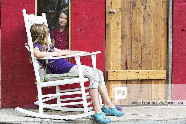 Ein junges Mädchen schaut aus dem Fenster auf ein anderes Mädchen  das in einem Schaukelstuhl auf der Veranda sitzt; Salmon Arm  British Columbia  Kanada .