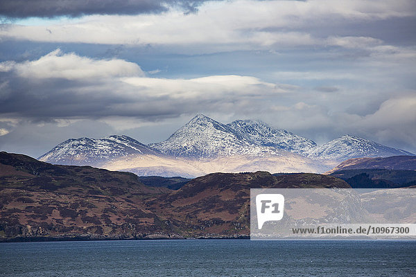 Hügel entlang der Küste und schneebedeckte Bergspitzen unter einem bewölkten Himmel; Argyll  Schottland'.