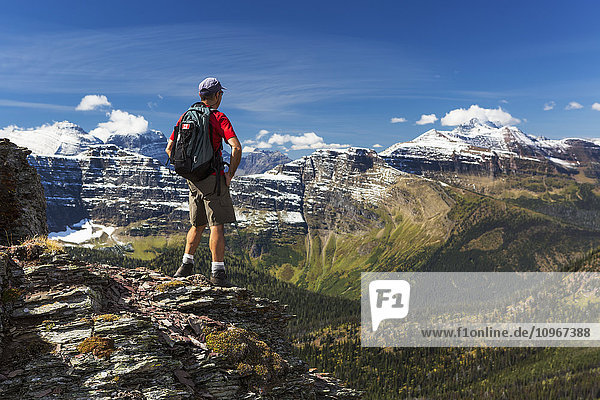 'Male hiker standing on top of mountain ridge overlooking snow peaked mountains and forest valley below with blue sky and clouds; Waterton Alberta Canada'