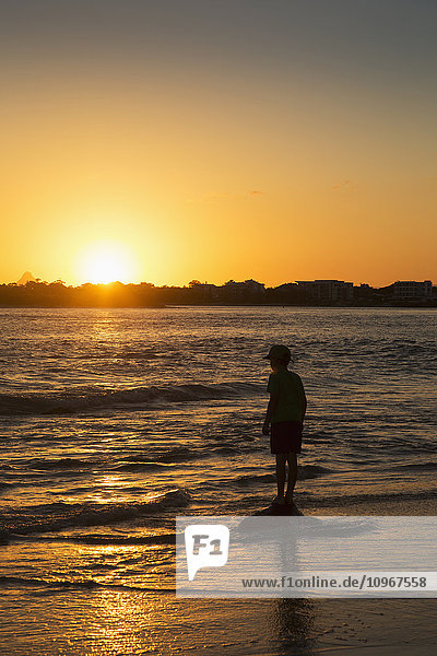 Silhouette eines kleinen Jungen  der bei Sonnenuntergang am Wasser steht; Caloundra  Queensland  Australien