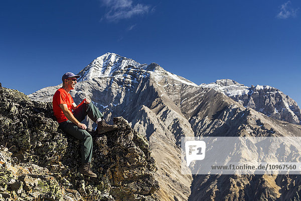 'Male hiker sitting on top of mountain ridge overlooking snow peaked mountains in the background with blue sky and clouds  Kananaskis Provincial Park; Alberta  Canada'
