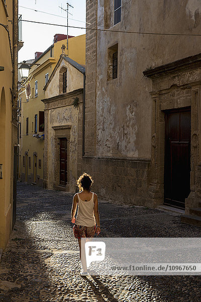 Mädchen  das eine kopfsteingepflasterte Gasse zwischen Gebäuden entlanggeht; Alghero  Sardinien  Italien'.