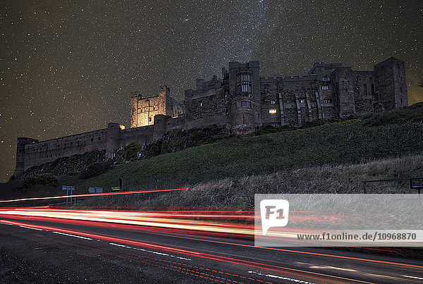 Lichtspuren auf einer Straße und ein sternenübersäter Himmel über Bamburgh Castle bei Nacht; Bamburgh  Northumberland  England'.