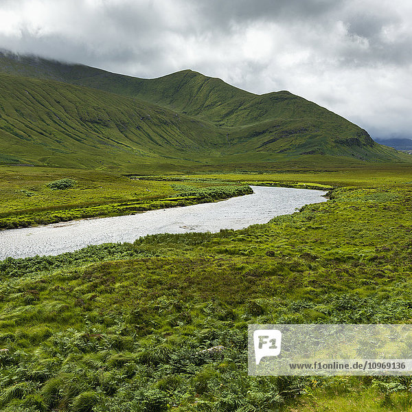 Berge und mit grünem Laub bedeckte Felder  durch die ein Fluss fließt  unter einem wolkenverhangenen Himmel in den Highlands; Schottland
