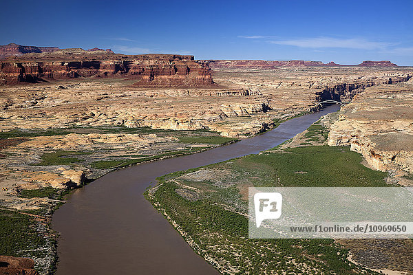 Luftaufnahme des Colorado River  der sich durch die Canyon-Landschaft im Süden Utahs schlängelt  mit einer Brücke  die den Fluss überspannt  im Hintergrund rote Canyonwände und blauer Himmel; Utah  Vereinigte Staaten von Amerika'.