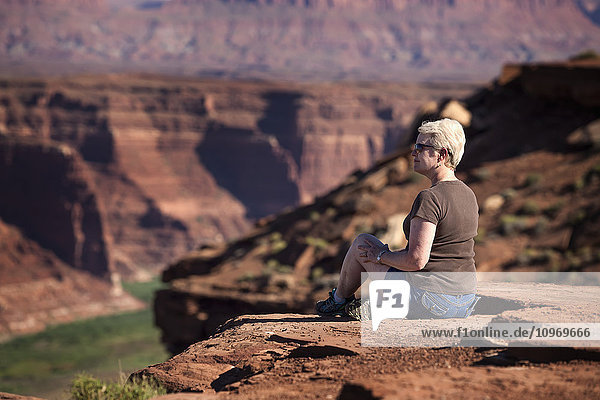 'Woman sitting on the edge of a rock canyon cliff looking over the Colorado river with canyon walls in the background; Colorado  United States of America'