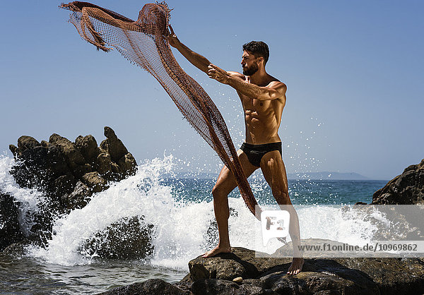 'A muscular man in a bathing suit throwing a net into the water while standing on a rock at the coast; Tarifa  Cadiz  Andalusia  Spain'