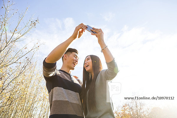 Ein junges asiatisches Paar genießt die gemeinsame Zeit in einem herbstlichen Park und macht Selbstporträts mit einem Smartphone in der späten Nachmittagssonne; Edmonton  Alberta  Kanada'.