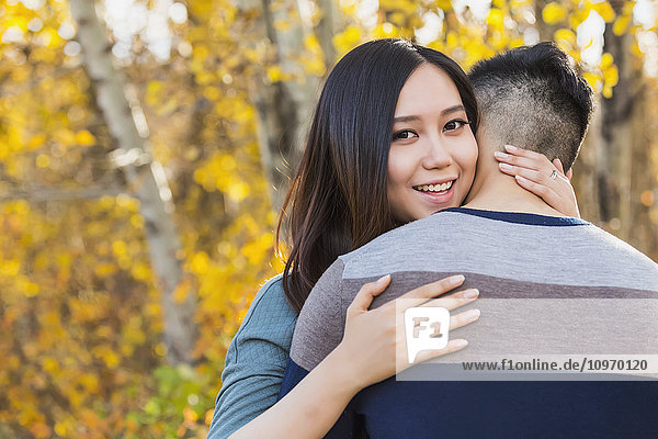 'A young Asian woman hugging her boyfriend in a park in autumn and looking over his shoulder at the camera; Edmonton  Alberta  Canada'