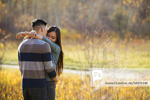 'A young Asian couple enjoying a romantic time together outdoors in a park in autumn and embracing each other in the warmth of the sunlight during the early evening; Edmonton  Alberta  Canada'