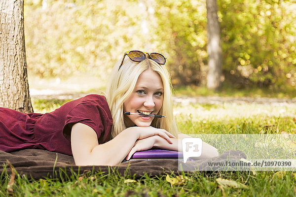 Eine junge Frau  die an einem warmen Herbsttag in einem Park Hausaufgaben macht und mit einem Stift im Mund für die Kamera posiert; Edmonton  Alberta  Kanada'.