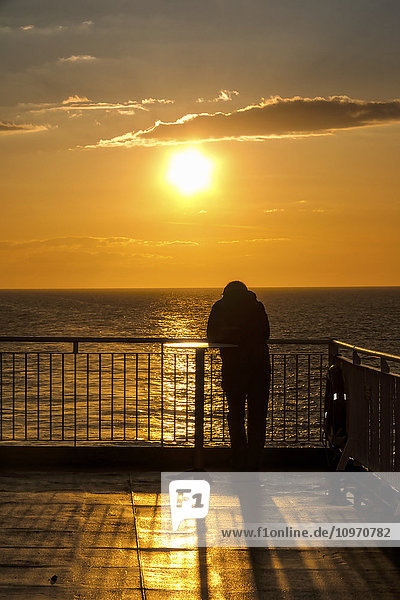 Ein Erwachsener steht an der Reling und blickt auf die Nordsee mit einem goldenen Sonnenuntergang; England'.
