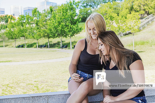 Zwei Freundinnen  die im Freien in einem Park gemeinsam auf ein Smartphone schauen; Edmonton  Alberta  Kanada'.