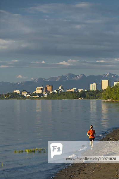 Mann beim Joggen am Strand neben dem Tony Knowles Coastal Trail während der Flut mit der Skyline der Stadt Anchorage  dem Knik Arm und den Chugach Mountains im Hintergrund; Anchorage  Alaska  Vereinigte Staaten von Amerika