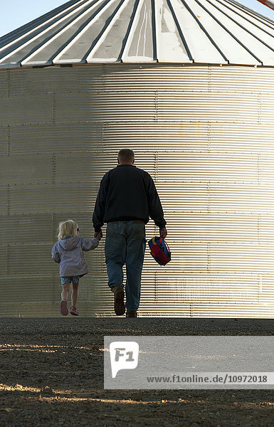 Agriculture - A farmer and his young daughter walk hand-in-hand through his farm yard with a grain bin in the background / near Sioux City  Iowa  USA.