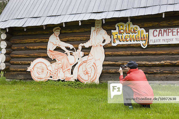 Woman tourist photographs a log cabin and motorcycle sign at the Tetsa River Lodge  British Columbia  Canada  Summer