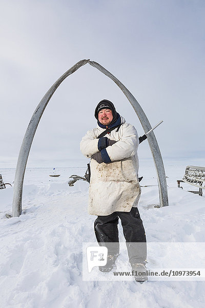 Ein einheimischer Jäger in Alaska  der einen weißen Kuspuk trägt und ein Gewehr in der Hand hält  steht unter dem Browerville-Walfischknochenbogen  Barrow  North Slope  Arktisches Alaska  USA  Winter'