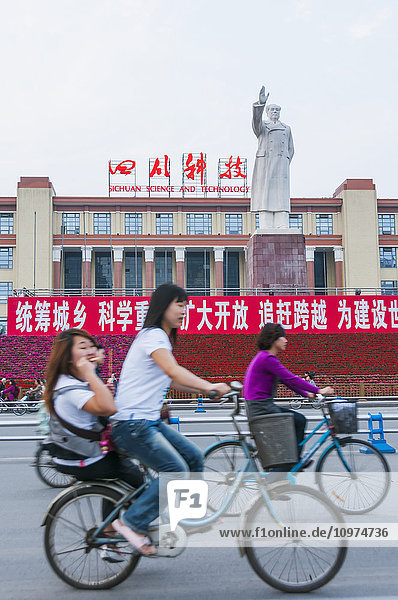'Chinese people riding bicycles on the street while Mao Zedong statue raises his hand; Chengdu  Sichuan province  China'