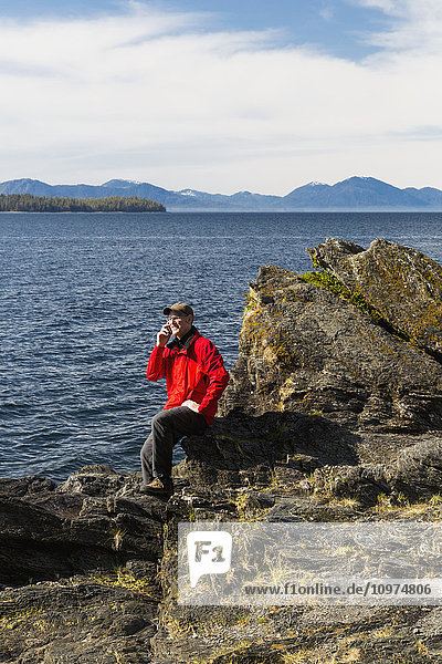 Ein Mann spricht mit seinem Smartphone am felsigen Ufer der Tongass Narrows  Ketchikan  Südost-Alaska  Frühling
