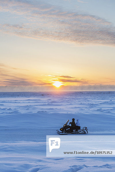 Zwei einheimische Alaskaner fahren mit einem Schneemobil durch eine schneebedeckte Landschaft bei Sonnenuntergang  Barrow  North Slope  Arctic Alaska  Winter