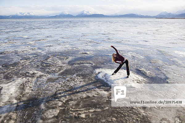 Frau dehnt sich auf einem Eisbrocken vor einem Lauf am Strand von Homer  Kenai-Halbinsel  Süd-Zentral-Alaska