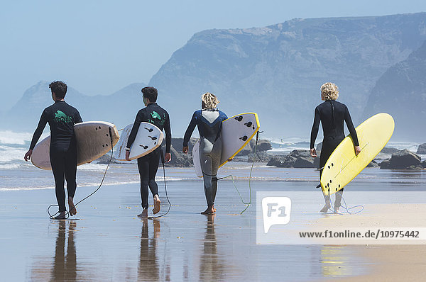 Junge männliche Surfer  die mit ihren Surfbrettern auf dem nassen Strand Richtung Wasser laufen; Praia da Cordoama  Portugal'.