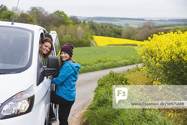 Zwei junge Frauen posieren vor ihrem Fahrzeug  während sie durch die englische Landschaft fahren  die mit leuchtend gelben Rapsblüten bedeckt ist; Cotswolds  England'.