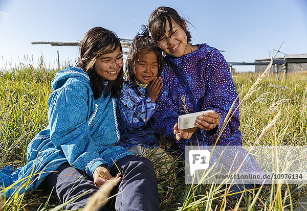 Athabascan-Mädchen in traditionellen Kuspuks  die im Gras sitzen und mit einem Smartphone telefonieren  mit einem Fischtrockengestell im Hintergrund  lächelnd  Nome  Alaska