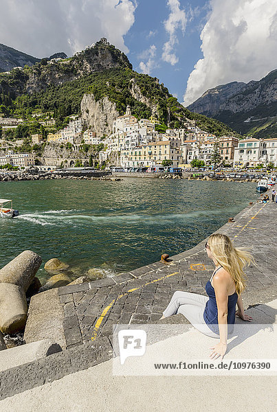 Eine junge Touristin posiert für ein Foto an der Strandpromenade mit der Stadt Amalfi im Hintergrund während eines Urlaubs in Italien; Amalfi  Provinz Salerno  Italien'.