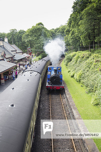 Dampf einer Lokomotive  die die Gleise hinunterfährt  während die Fahrgäste in einem Bahnhof auf das Einsteigen in einen Zug warten; Cumbria  England'.